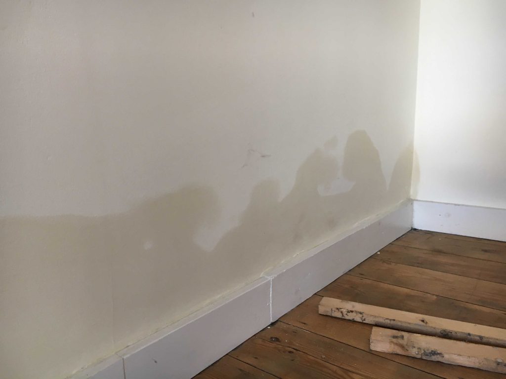 Corner of a room with a wooden floor and white wall showing water damage.